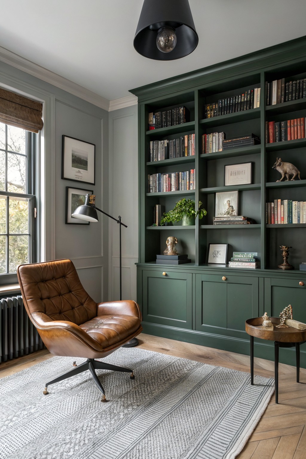 Cozy study room featuring deep green built-in bookcases and cabinets against light gray walls, tan leather armchair, wood floors, and assorted books and decor