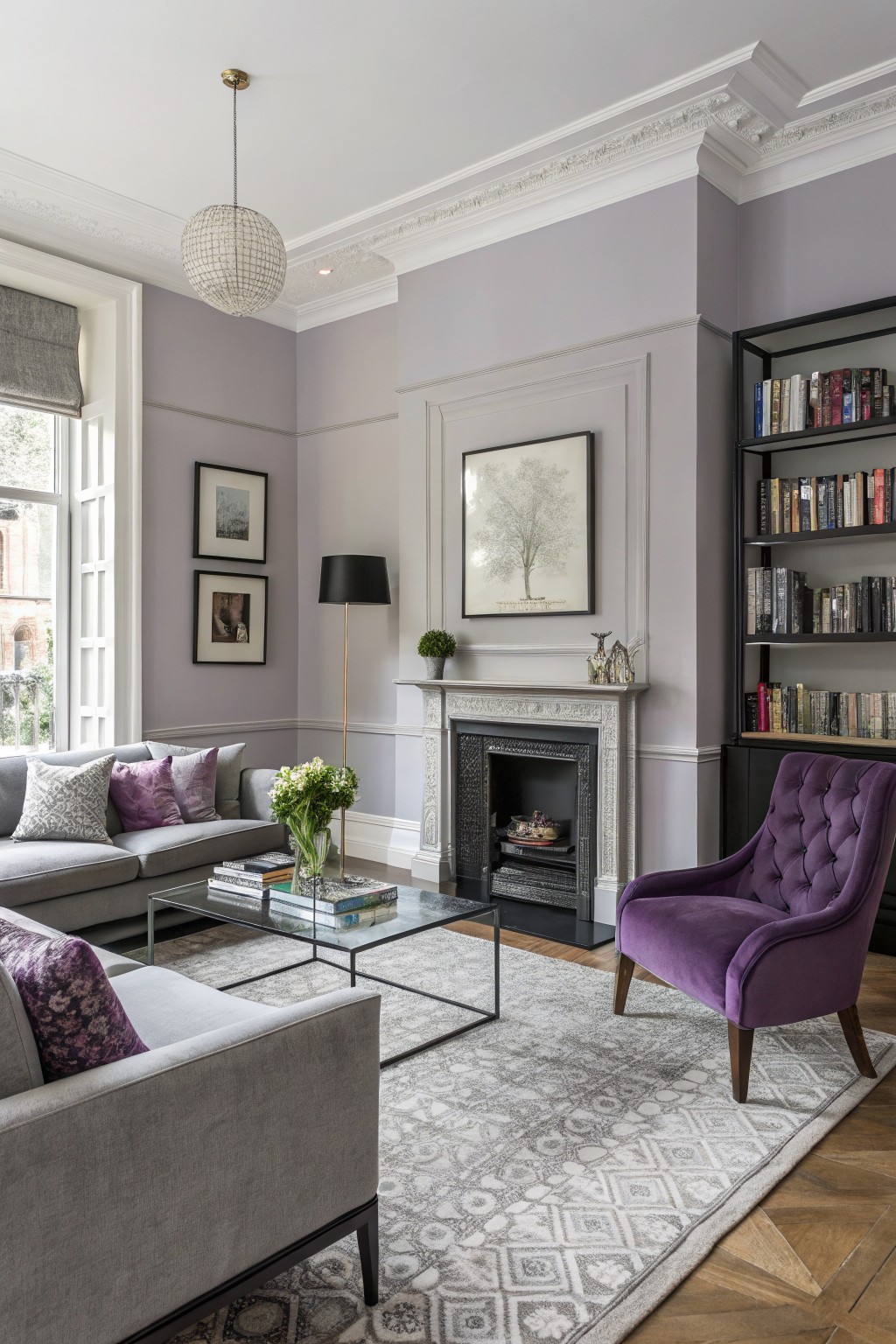 Living room with pale lavender-gray walls, gray sofa with purple pillows, purple velvet chair, glass coffee table, bookshelves, and ornate fireplace
