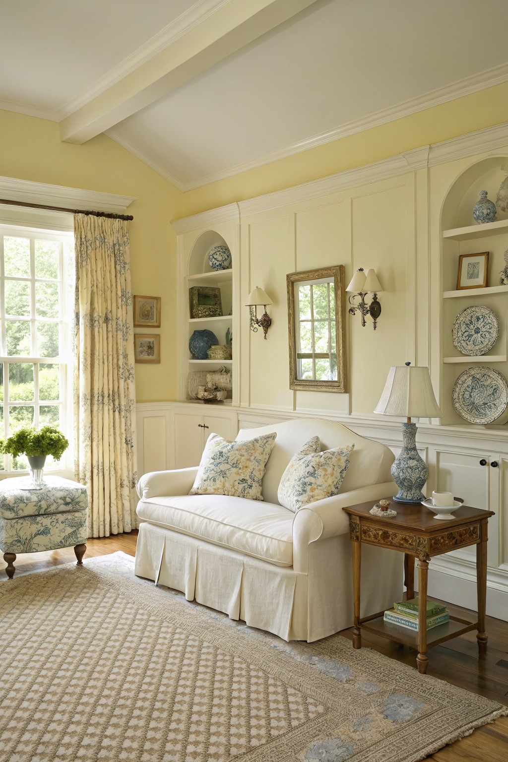 Cozy living room with pale yellow walls, white floral sofa, built-in shelves with blue and white china, and hardwood floors