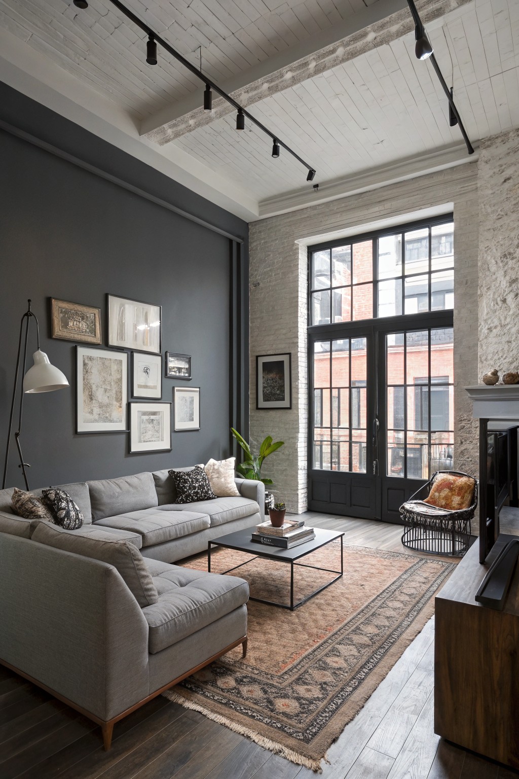 Loft-style living room featuring a dark gray accent wall, gray sectional sofa, large industrial windows, and exposed white brick ceiling