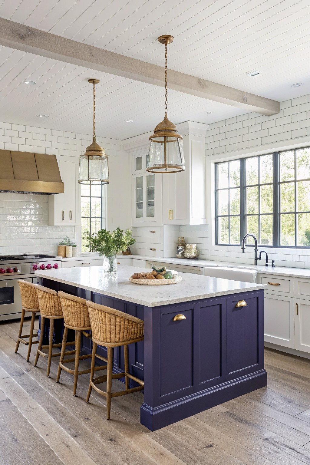 Modern kitchen featuring a deep navy island with white quartz countertop, rattan barstools, brass pendant lights, and large windows overlooking greenery