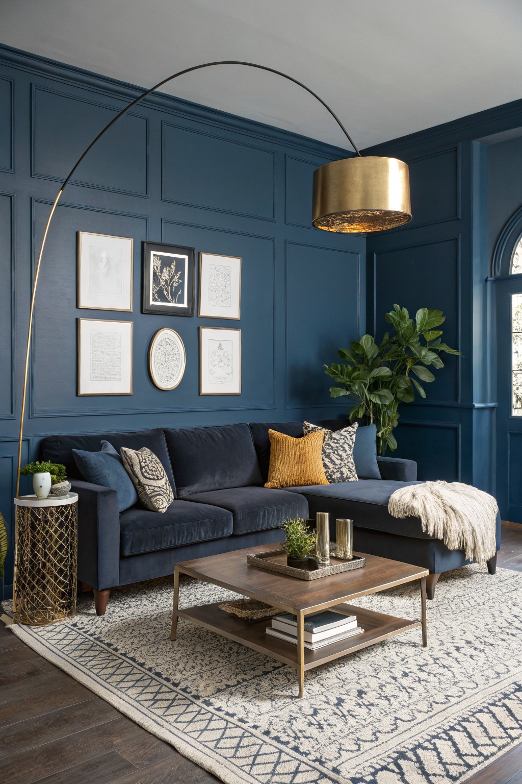 Living room featuring deep navy paneled walls, navy velvet sofa with mustard pillows, gold arc lamp, brass side table, and potted plants on hardwood floors