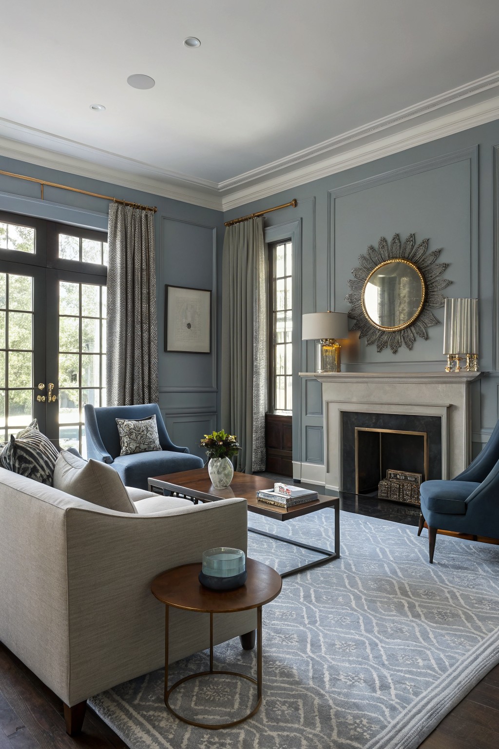 Living room with soft blue-gray paneled walls, beige sofa, blue accent chairs, wood coffee table, fireplace mantel, brass side table, and large windows.