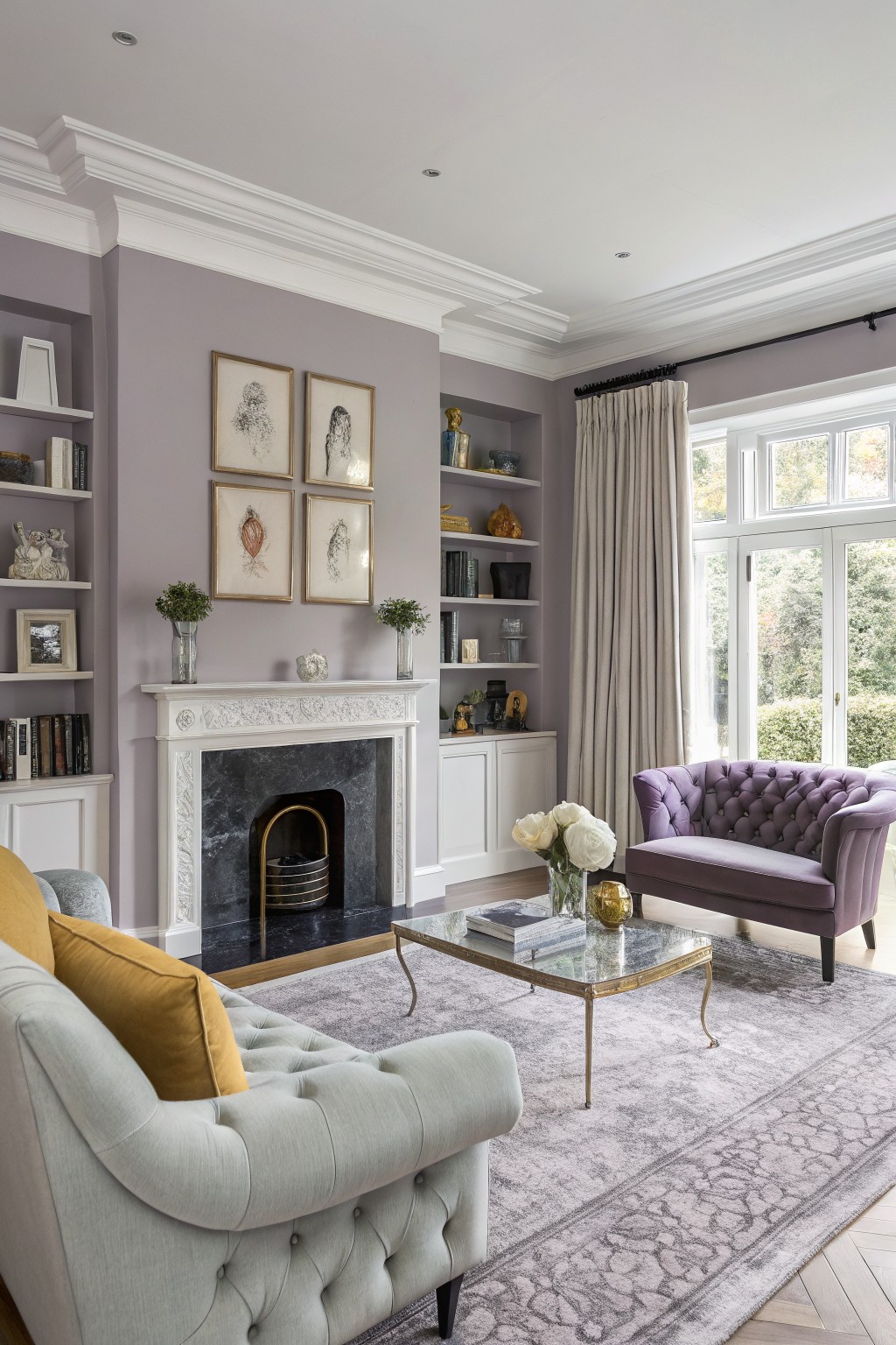 Living room with pale lilac gray walls, ornate white fireplace, light gray tufted sofa with yellow pillows, purple velvet chair, and gold-accented glass table on gray rug