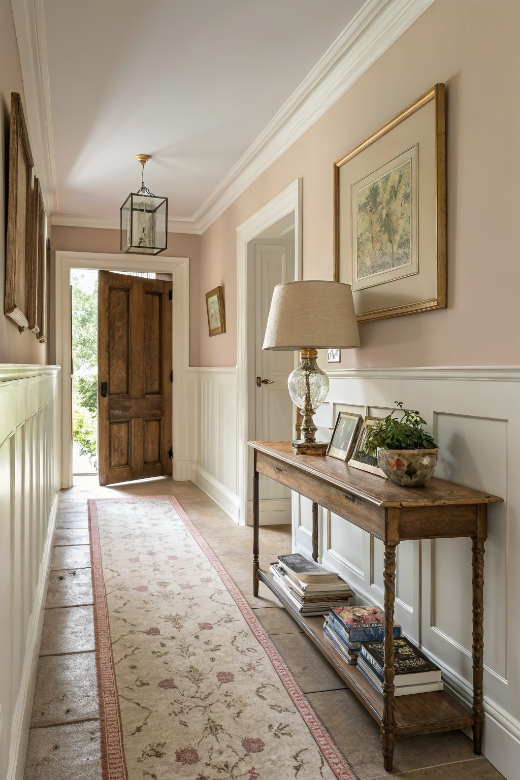 Hallway with pale greige upper walls, white wainscoting, wooden console table with lamp and books, and open front door to outdoors