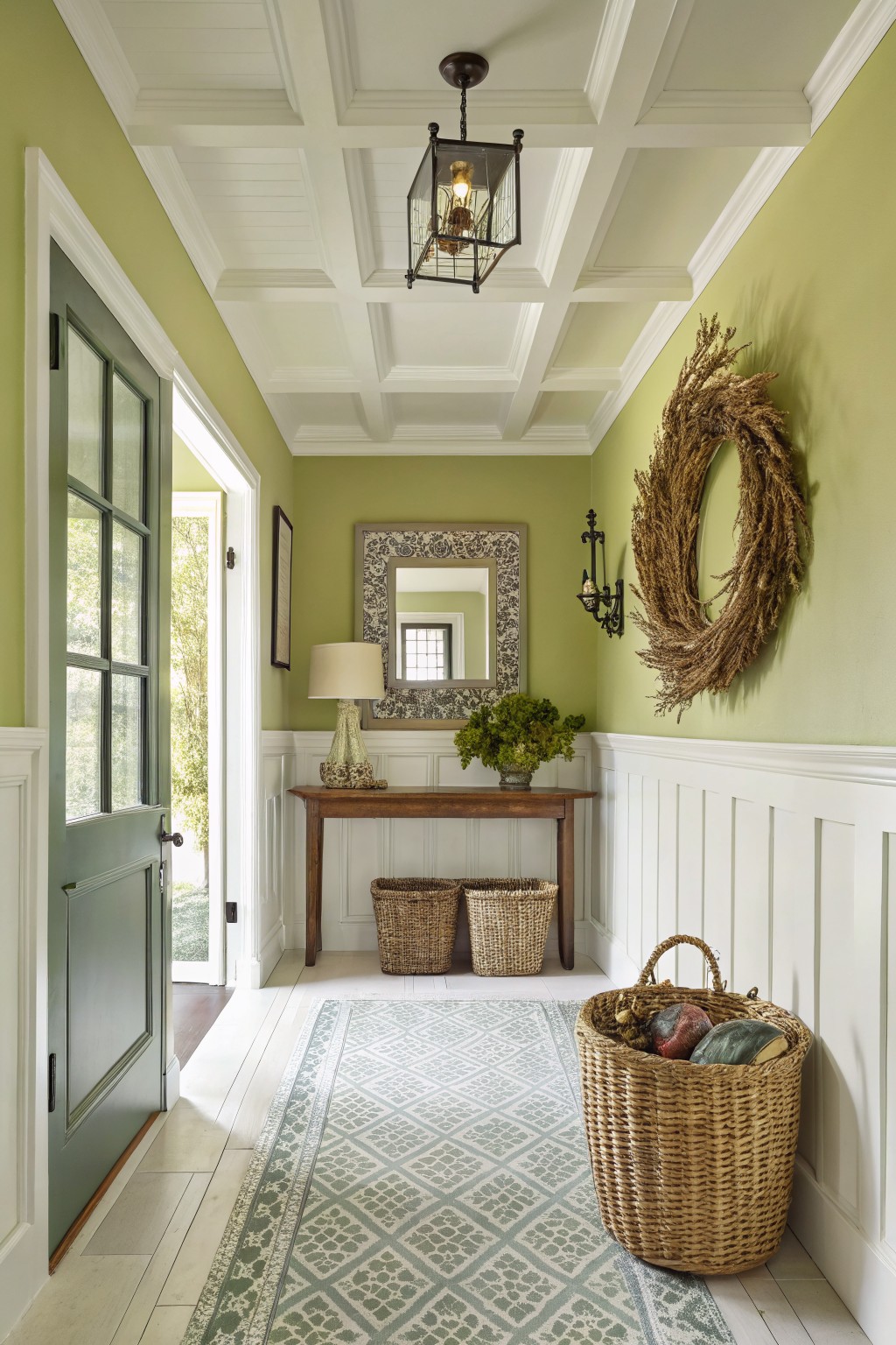 Pale sage green walls in a cozy entryway with white wainscoting, wooden console table holding a lamp and greenery, hanging wheat wreath, and baskets on the floor