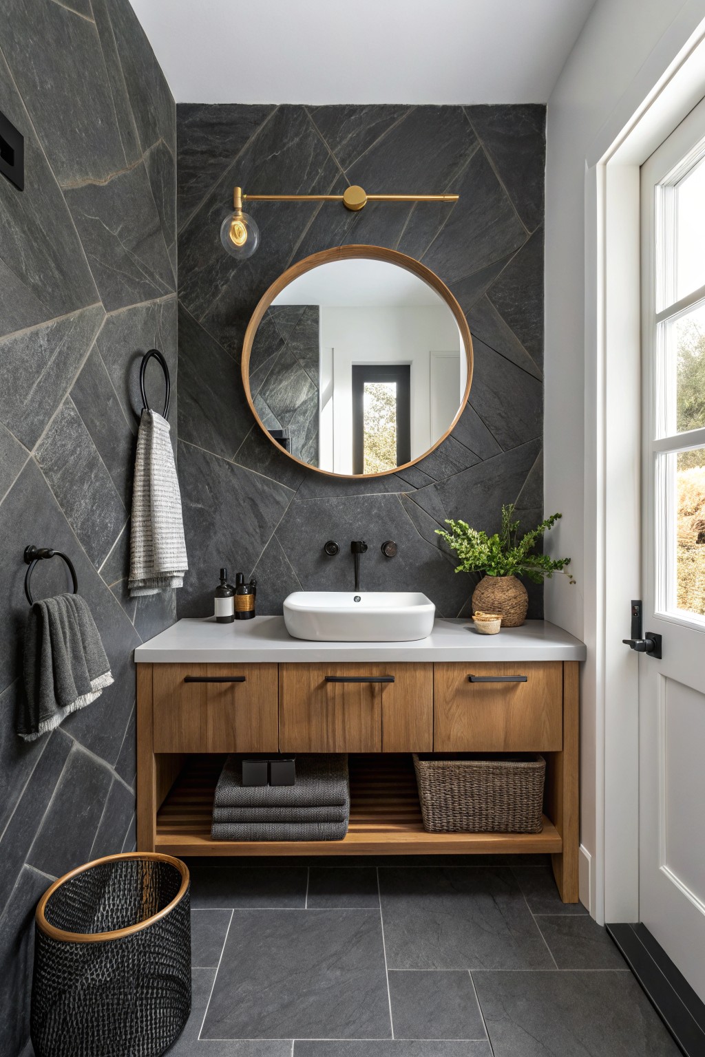 Modern powder room featuring slate gray tiled walls, oak vanity with woven baskets, round wood-framed mirror, gold sconce, and potted plants beside a white vessel sink