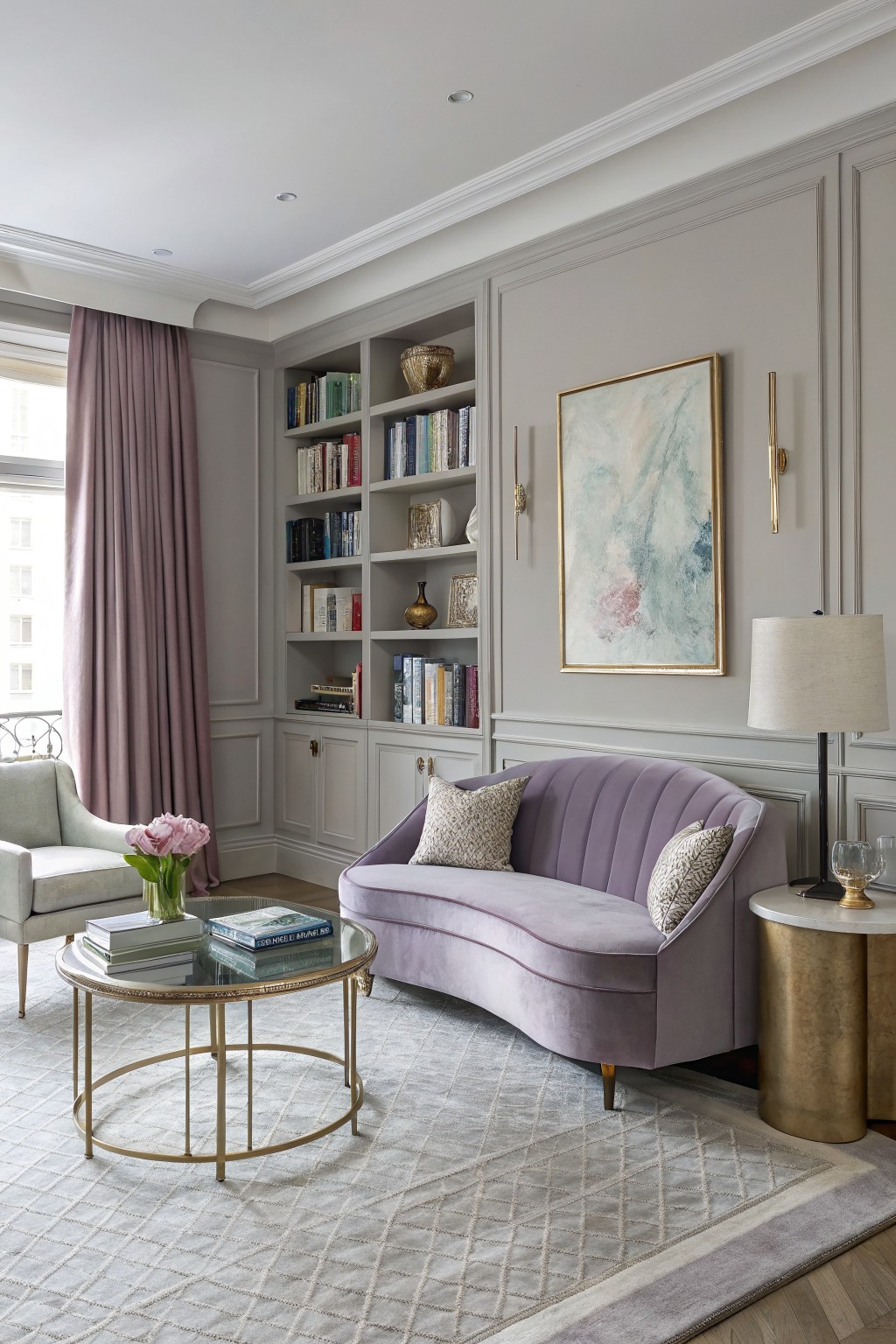 Open-concept living room featuring soft greige paneled walls, built-in bookshelves, purple velvet settee, gold coffee table, and large window with mauve drapes