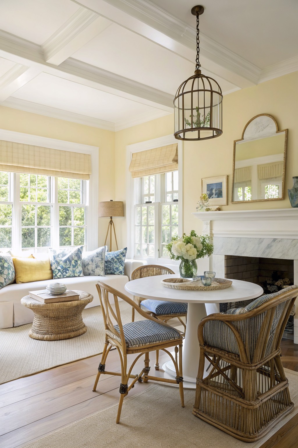Open concept living room with soft pale yellow walls, white trim, large windows, rattan chairs, round white table, and cozy sofa by marble fireplace