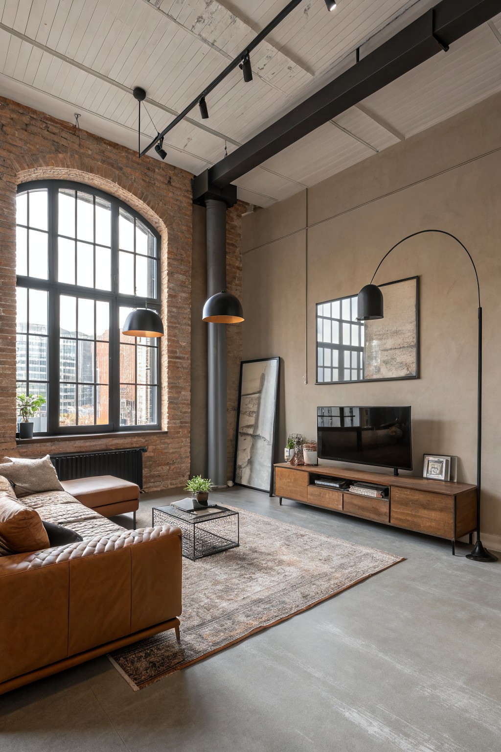Modern loft living room featuring warm greige walls with exposed brick accent, wooden TV console, tan leather sofa, and track lighting
