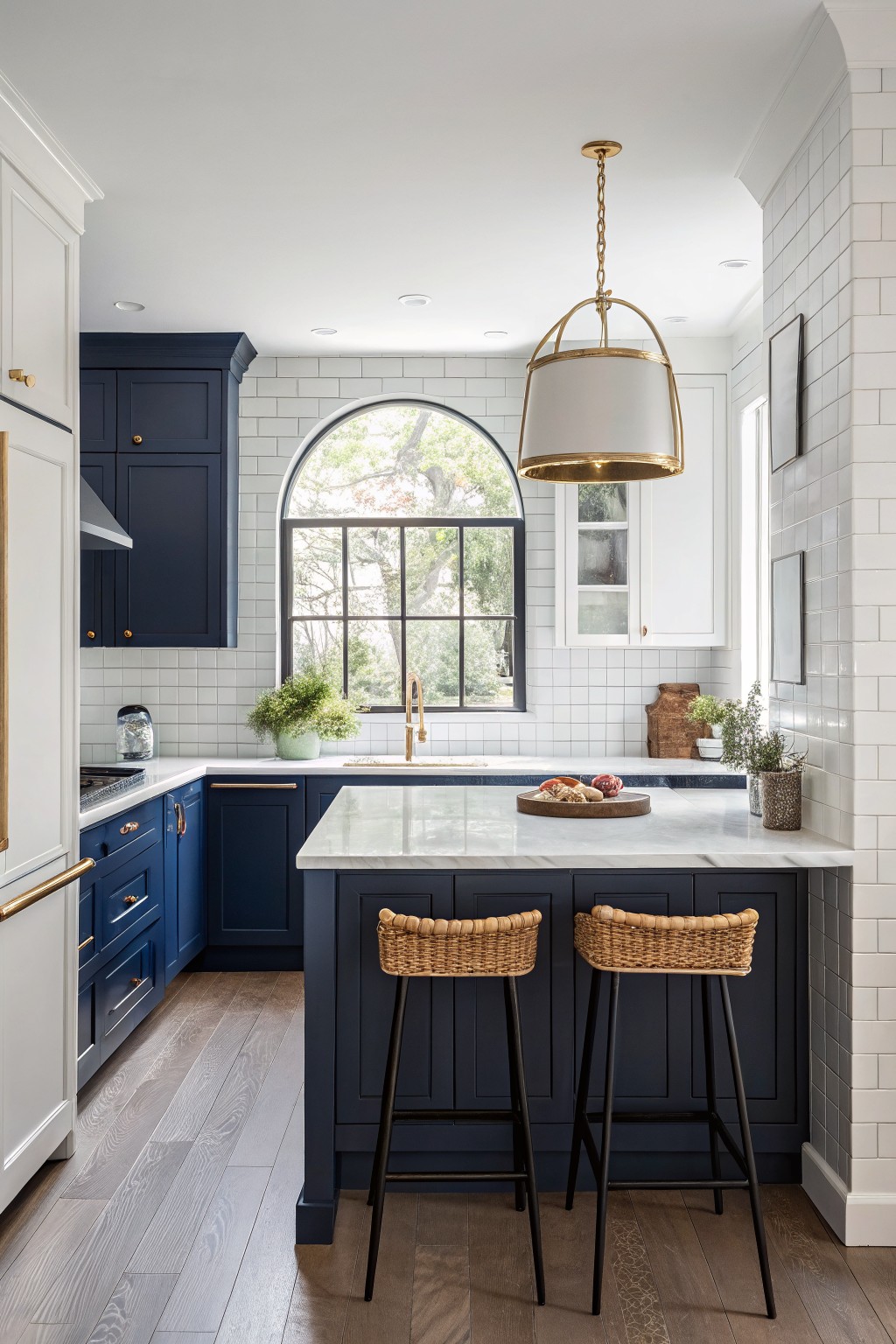 Modern kitchen featuring deep navy cabinets, white subway tile backsplash, marble counters, rattan stools, and an arched black-framed window overlooking trees
