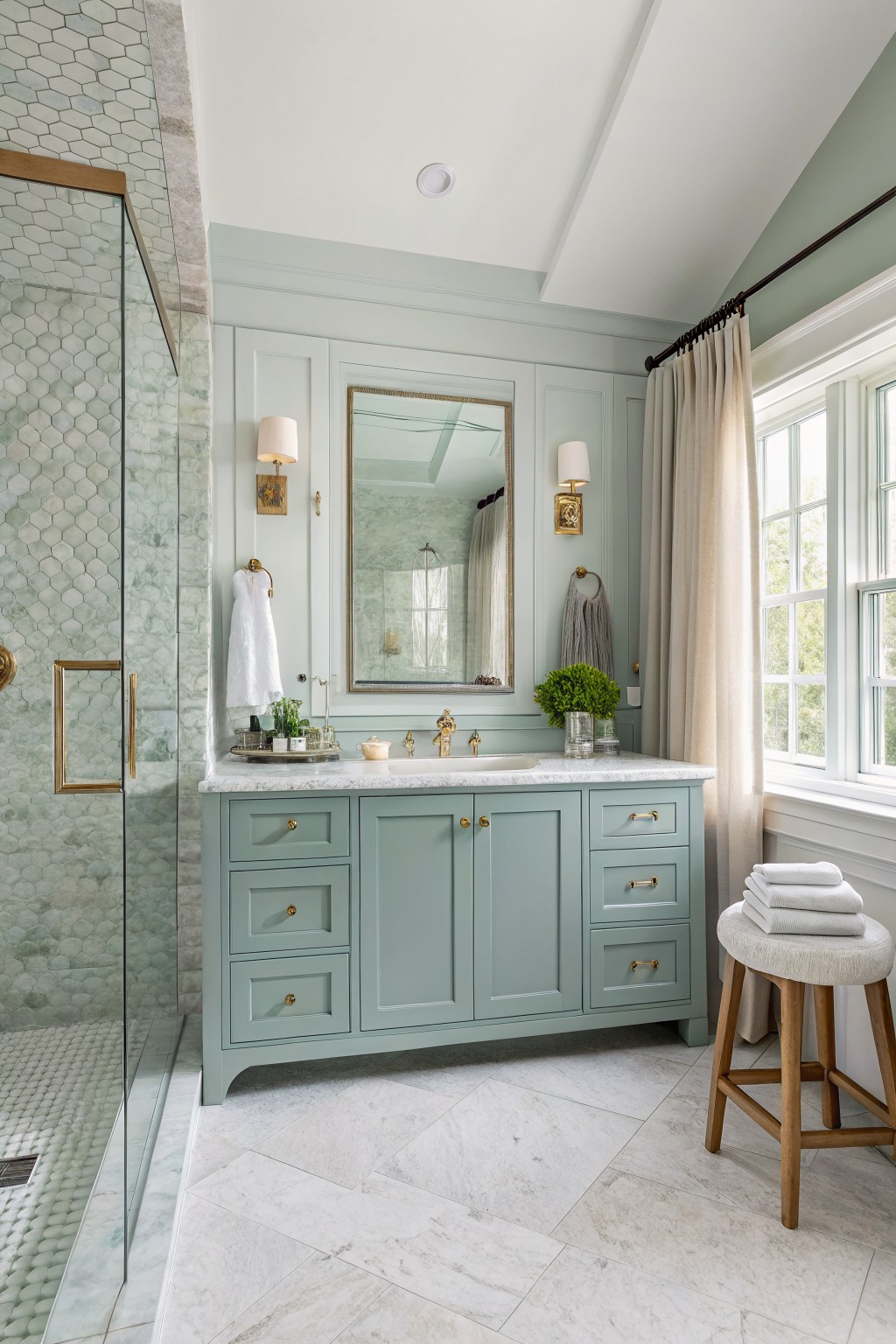 Modern bathroom featuring pale sage green painted walls and cabinets, gold fixtures, glass shower with green tile, marble floor, wood stool, and window with linen curtains