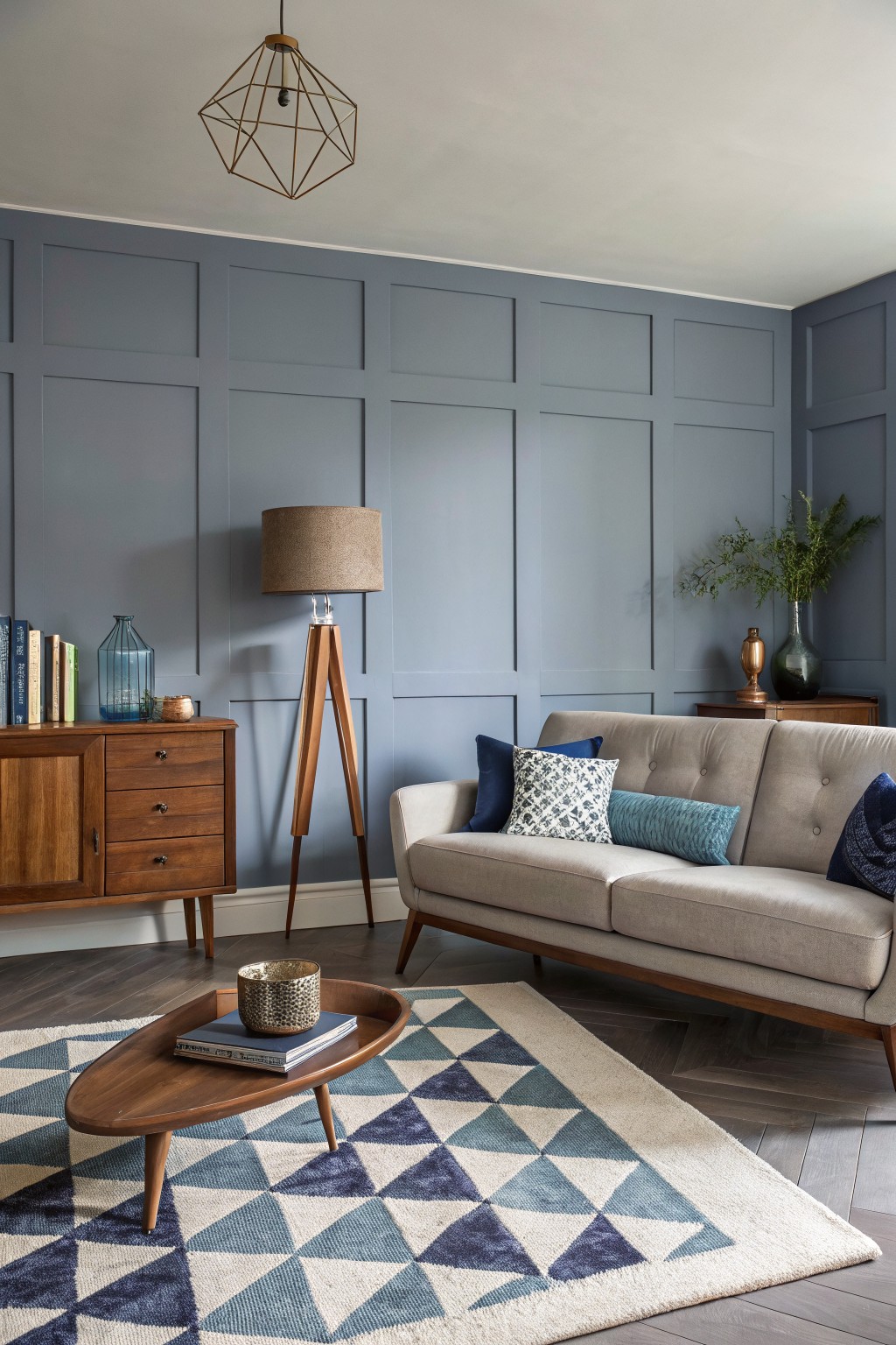 Living room with soft blue-gray paneled walls, wood sideboard, tripod floor lamp, gray sofa with patterned pillows, and geometric blue rug