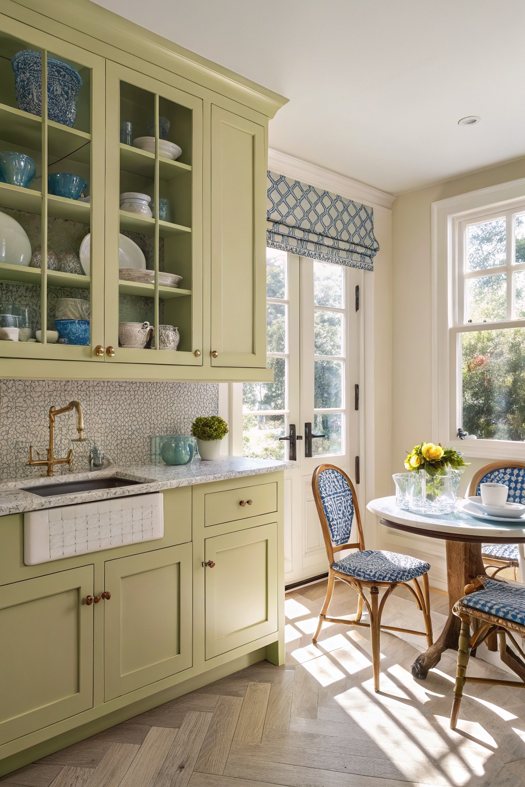 Kitchen with pale sage green cabinets, glass-front uppers holding blue and white dishes, brass hardware, white farmhouse sink, and rattan chairs by French doors letting in sunlight