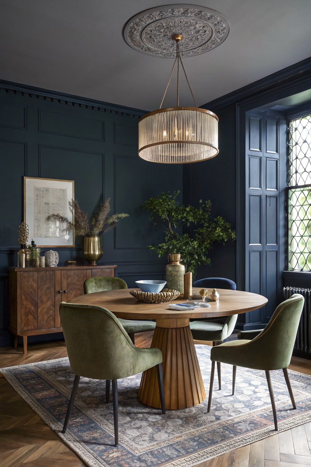 Dining room featuring deep navy paneled walls, round walnut pedestal table, green velvet chairs, brass chandelier, and potted plants on wood sideboard
