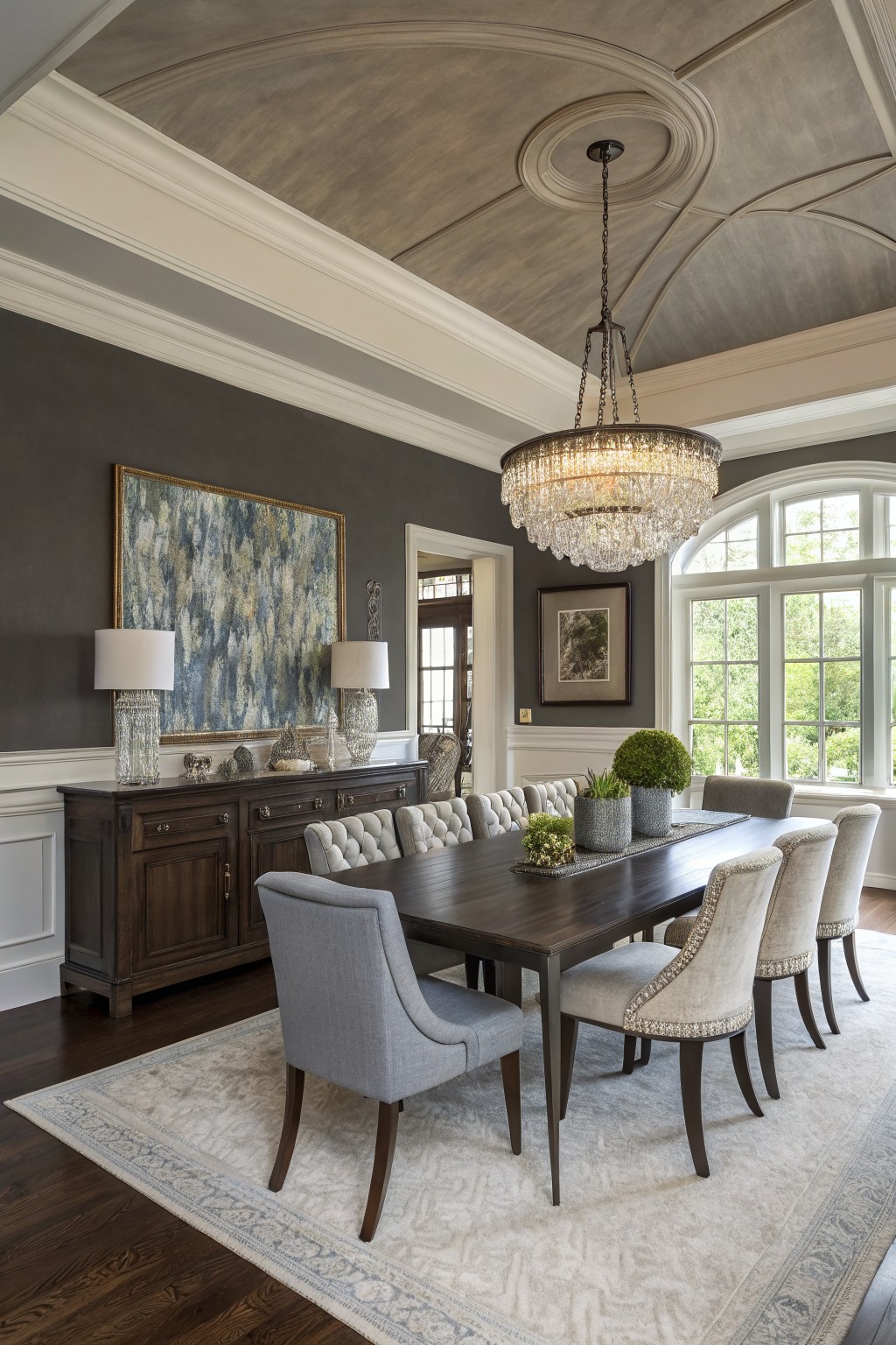 Dining room featuring warm gray walls, dark wood table and buffet, tufted chairs, crystal chandelier, and large abstract painting