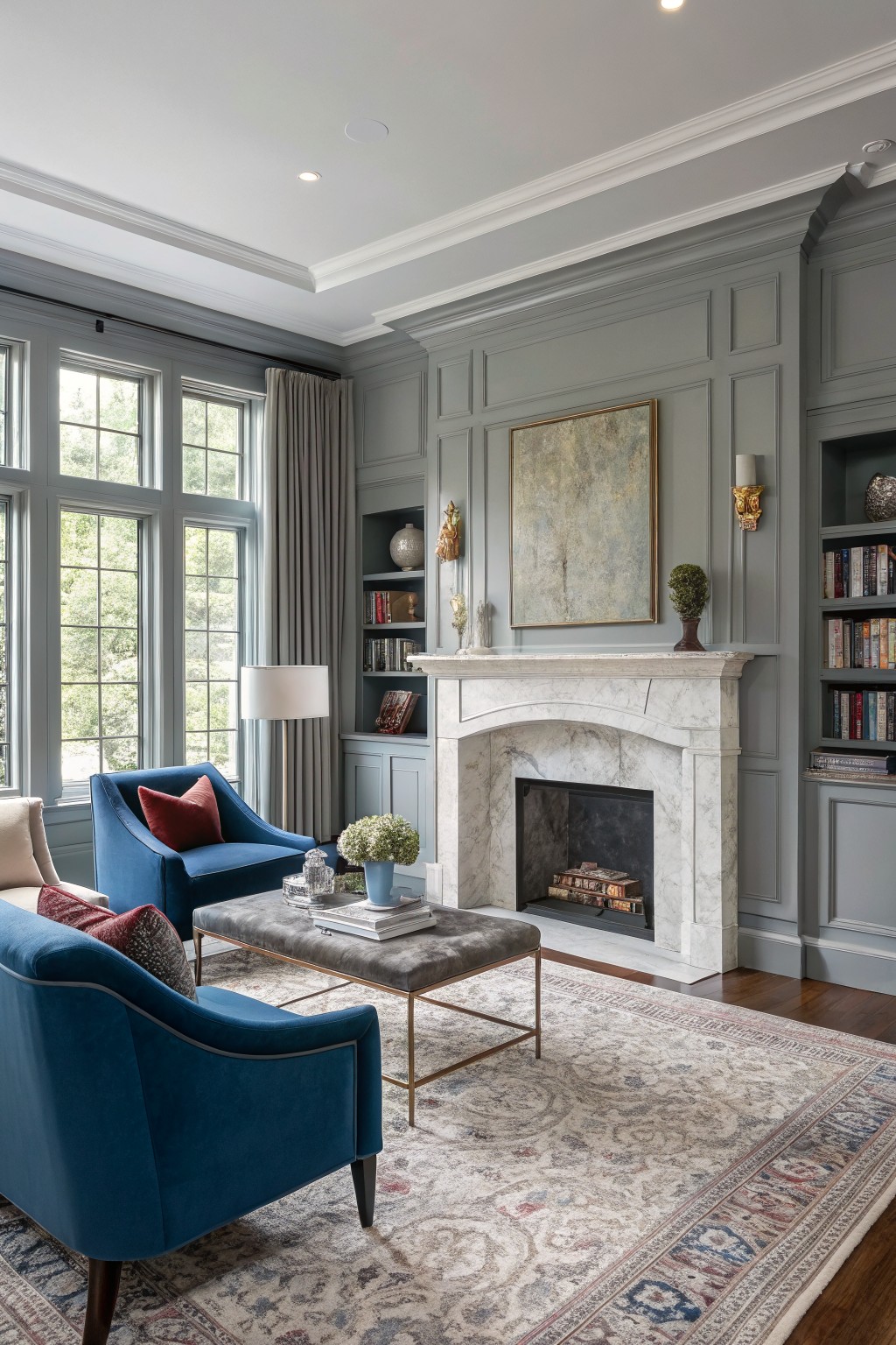 Cozy living room with soft gray paneled walls, marble fireplace, blue velvet chairs, bookshelves, and large windows overlooking trees