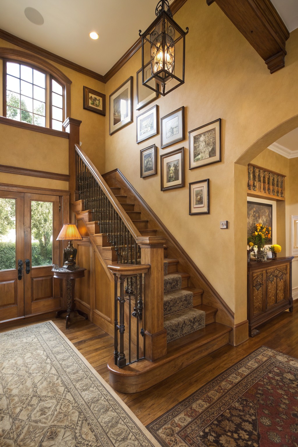 Grand two-story entry with warm beige walls, dark wood staircase and trim, wrought iron railing, wall art, and wooden side tables