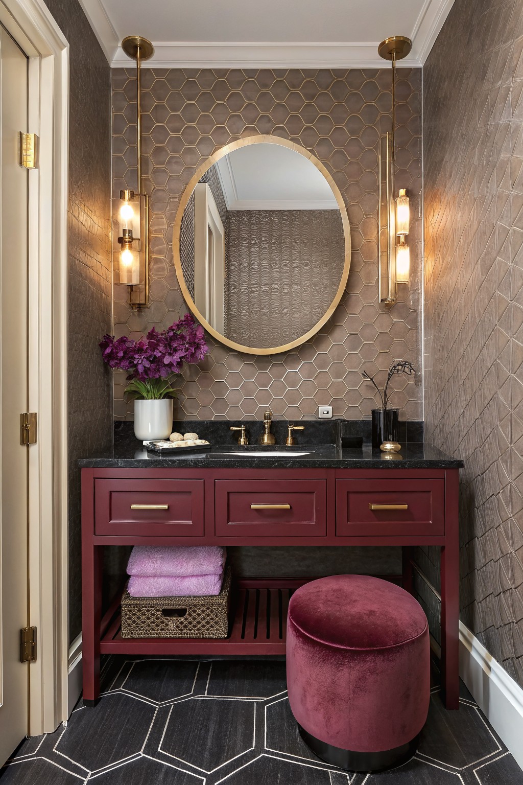 Small powder room featuring deep red painted vanity with gold pulls, large round mirror, metallic honeycomb wall tiles, purple flowers, and black hexagon floor tiles