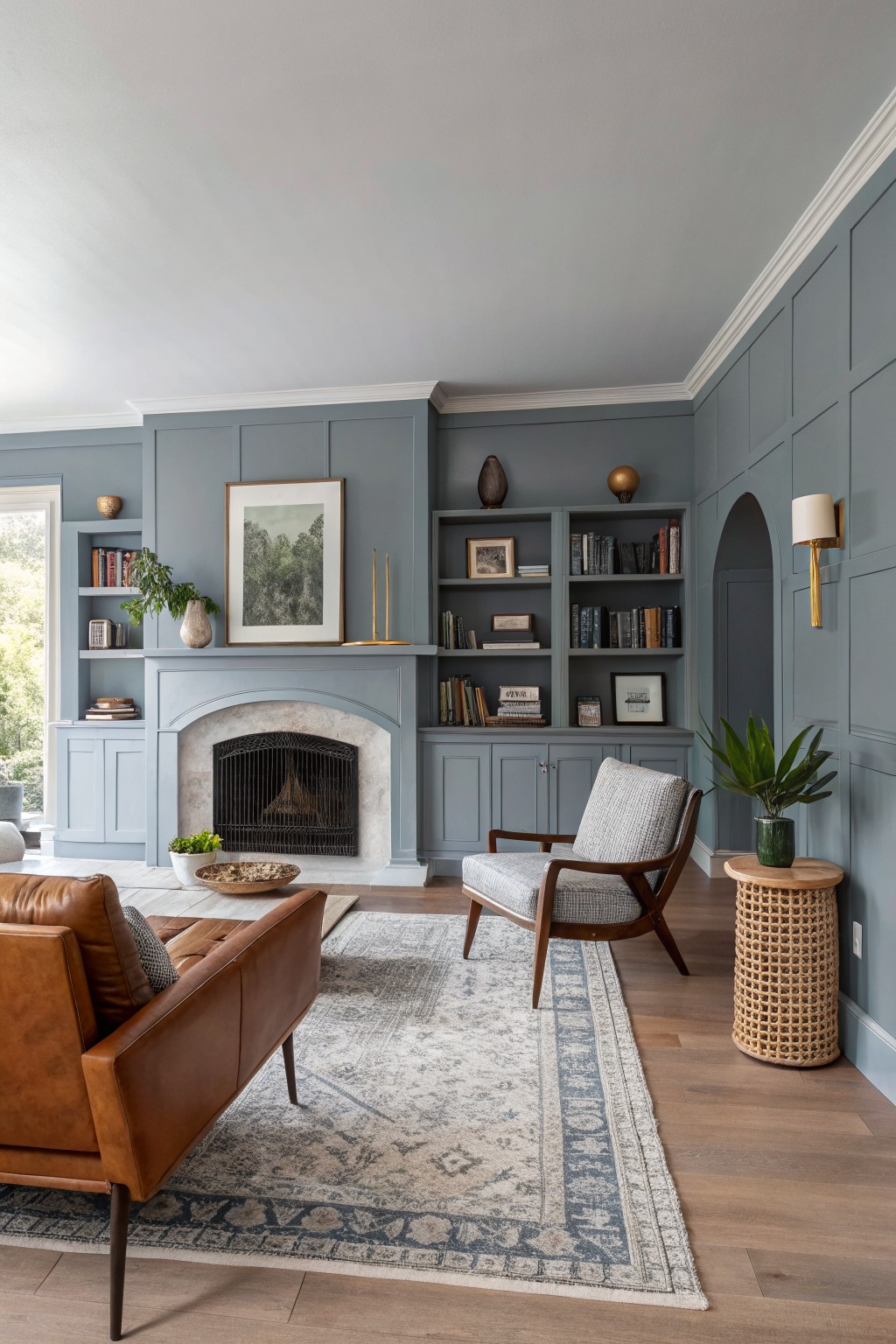 Cozy living room with soft blue-gray paneled walls, arched fireplace, built-in bookshelves, tan leather sofa, gray armchair, wood floors, and houseplants
