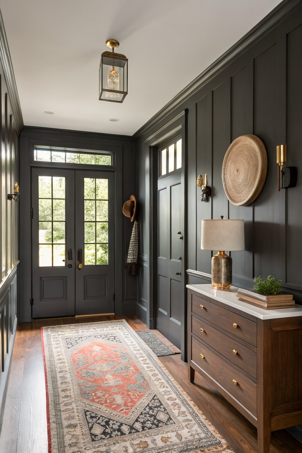 Narrow entry hallway with dark charcoal gray board and batten walls, oak wood floors, Persian-style runner rug, wood console table with lamp, and double doors to outdoors