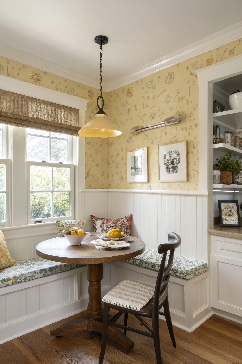 Cozy kitchen nook with crisp white beadboard wainscoting, pale yellow floral wallpaper, round wood pedestal table, and a window overlooking trees