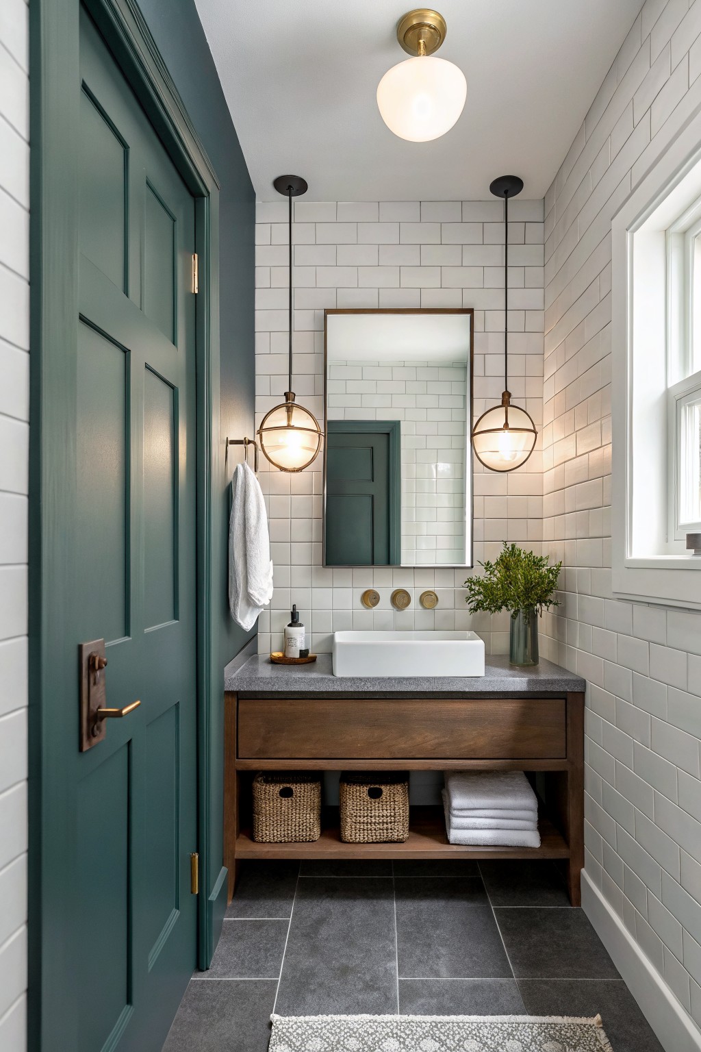 Small bathroom featuring a deep teal-green paneled door next to white subway tile walls, wood vanity with open storage, and brass pendant lights.