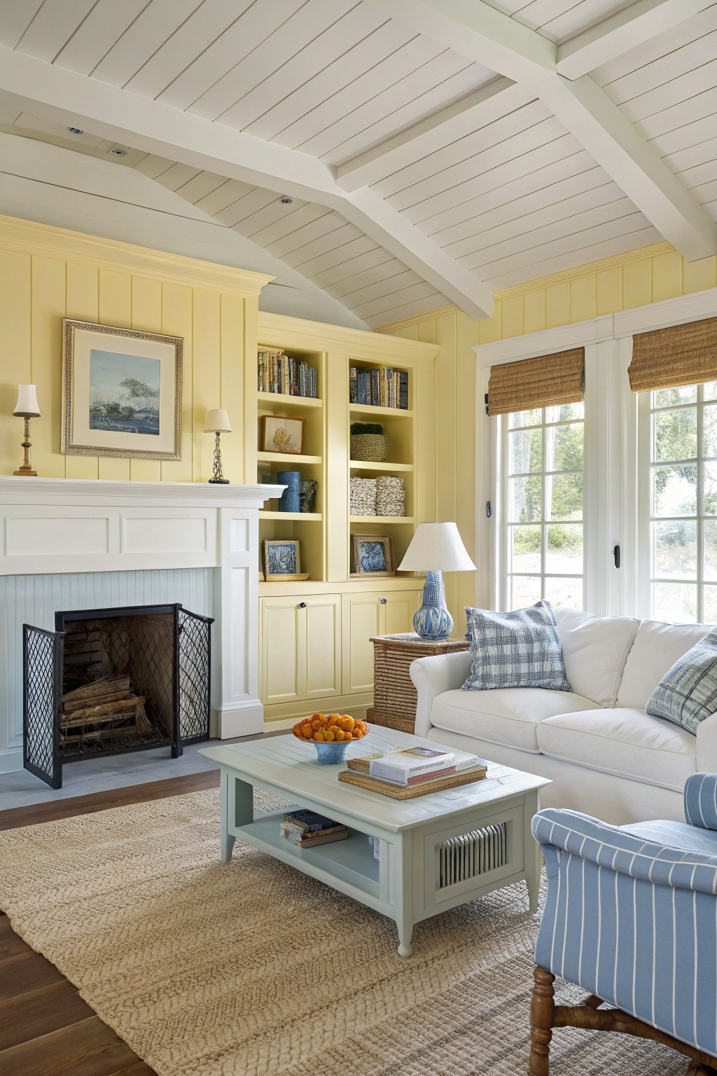 Living room with pale yellow paneled walls, white trim and ceiling beams, fireplace, white sofa with blue pillows, coffee table, and large windows with bamboo shades