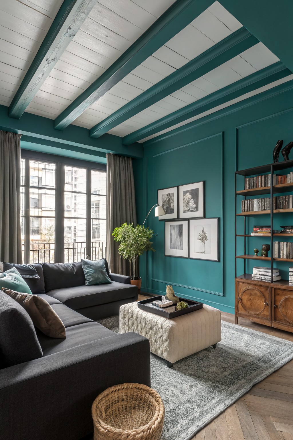 Cozy living room featuring deep teal walls and beams, dark gray sofa, wooden credenza, black metal bookshelves, and large windows with gray drapes