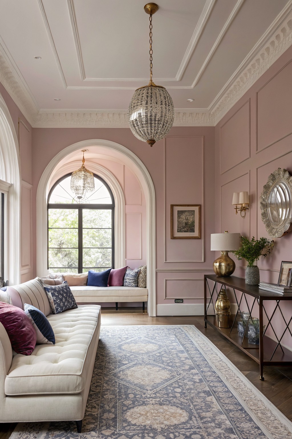 Living room with soft blush pink paneled walls, cream tufted sofa piled with pillows, brass console table, and arched window