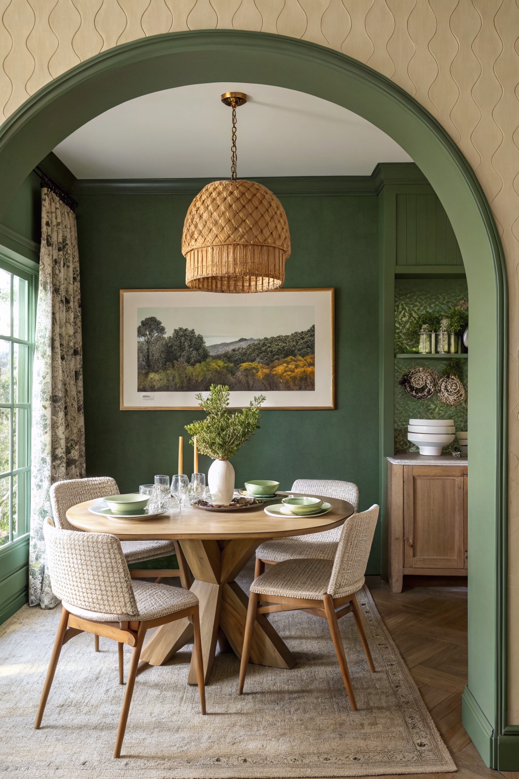 Dining room with deep green walls and curved archway framing a round wooden table set for dinner, rattan chairs, woven pendant light, and landscape artwork.