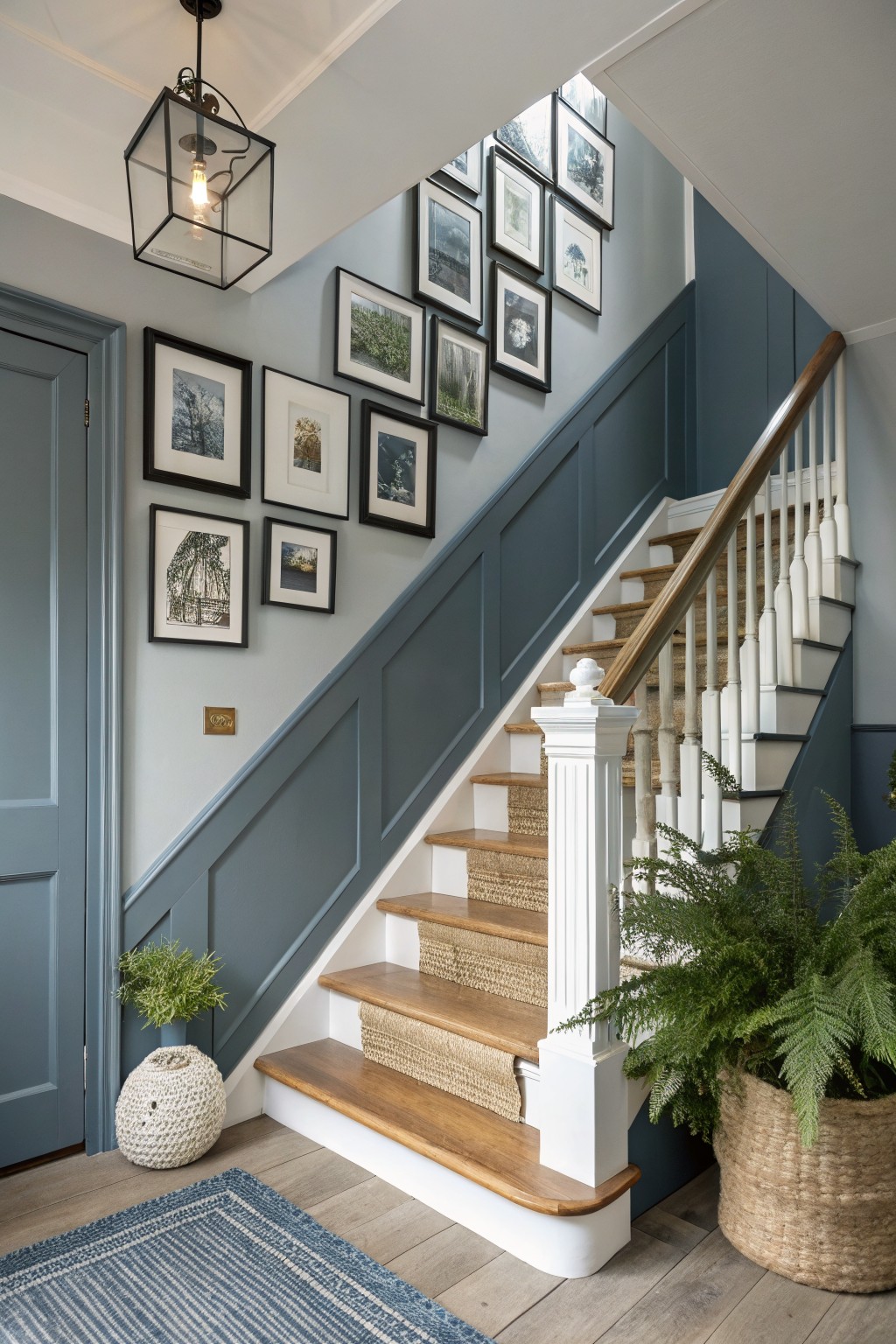 Stairwell with soft blue-gray painted wainscoting along white-trimmed stairs, oak treads covered in seagrass, potted ferns, woven basket, and black-framed art gallery wall.