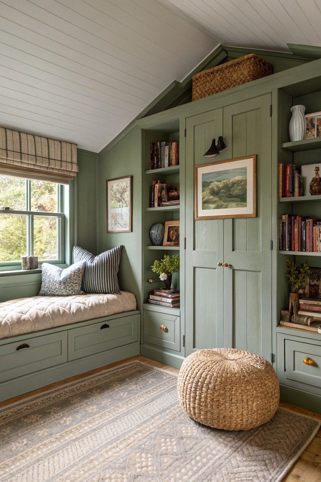 Cozy farmhouse bedroom reading nook with soft sage green built-in cabinetry, window seat cushions, bookshelves, and woven pouf on a patterned rug