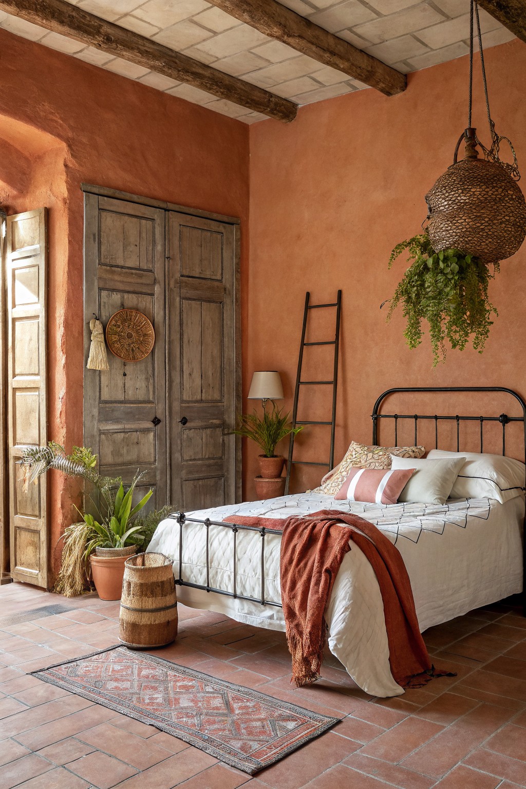 Farmhouse bedroom with warm terracotta walls, black metal bed frame, distressed wood wardrobe, potted greenery, and woven basket accents on terracotta tile floor