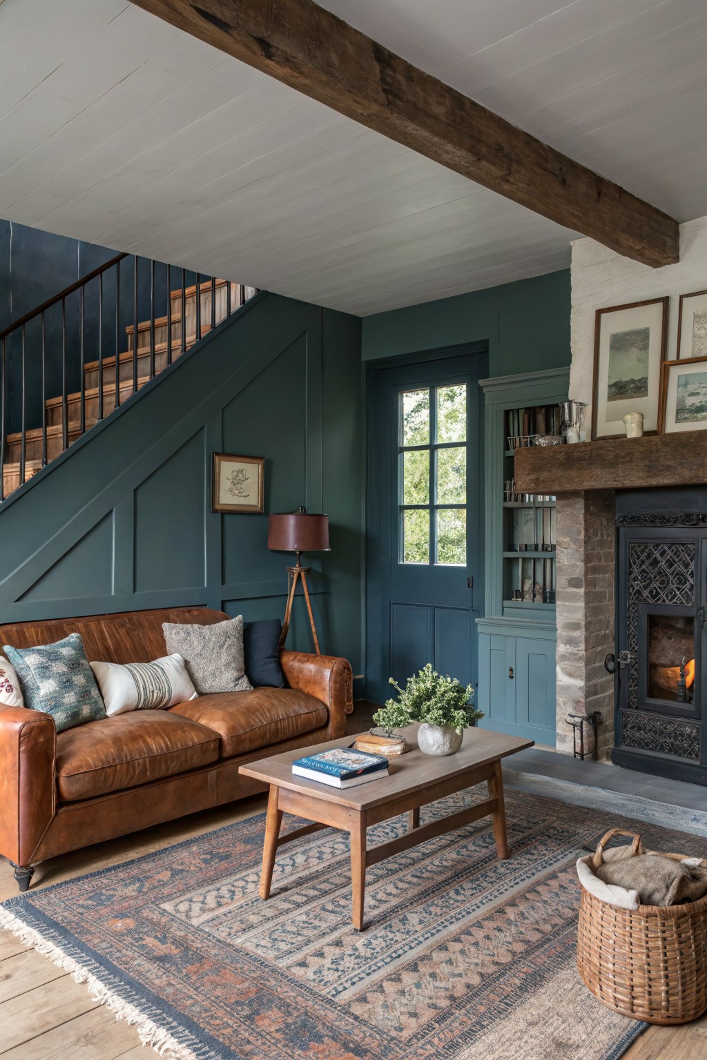 Living room with deep teal paneled walls, brown leather sofa by a stone fireplace, wood beams overhead, and a woven rug