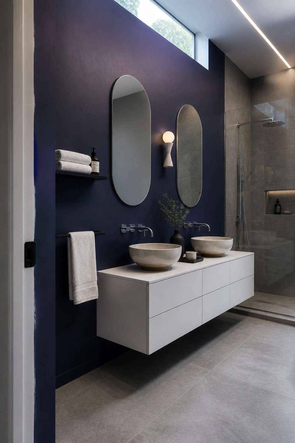 Modern bathroom with navy wall, white floating vanity, two stone vessel sinks, oval mirrors.