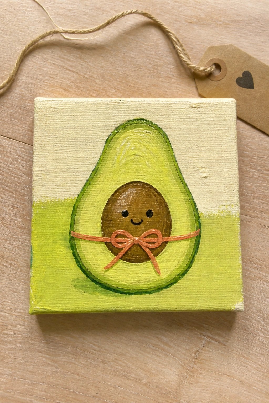 Small square canvas with a painted avocado half, smiling pit, orange bow, and light green background with attached tag.