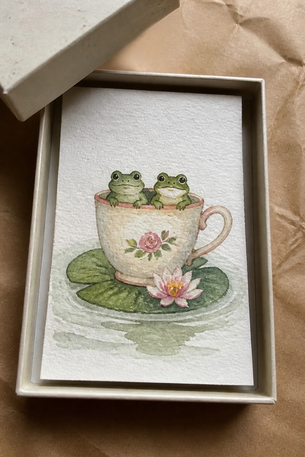 Watercolor painting of two frogs in floral teacup on lily pads with pink flower