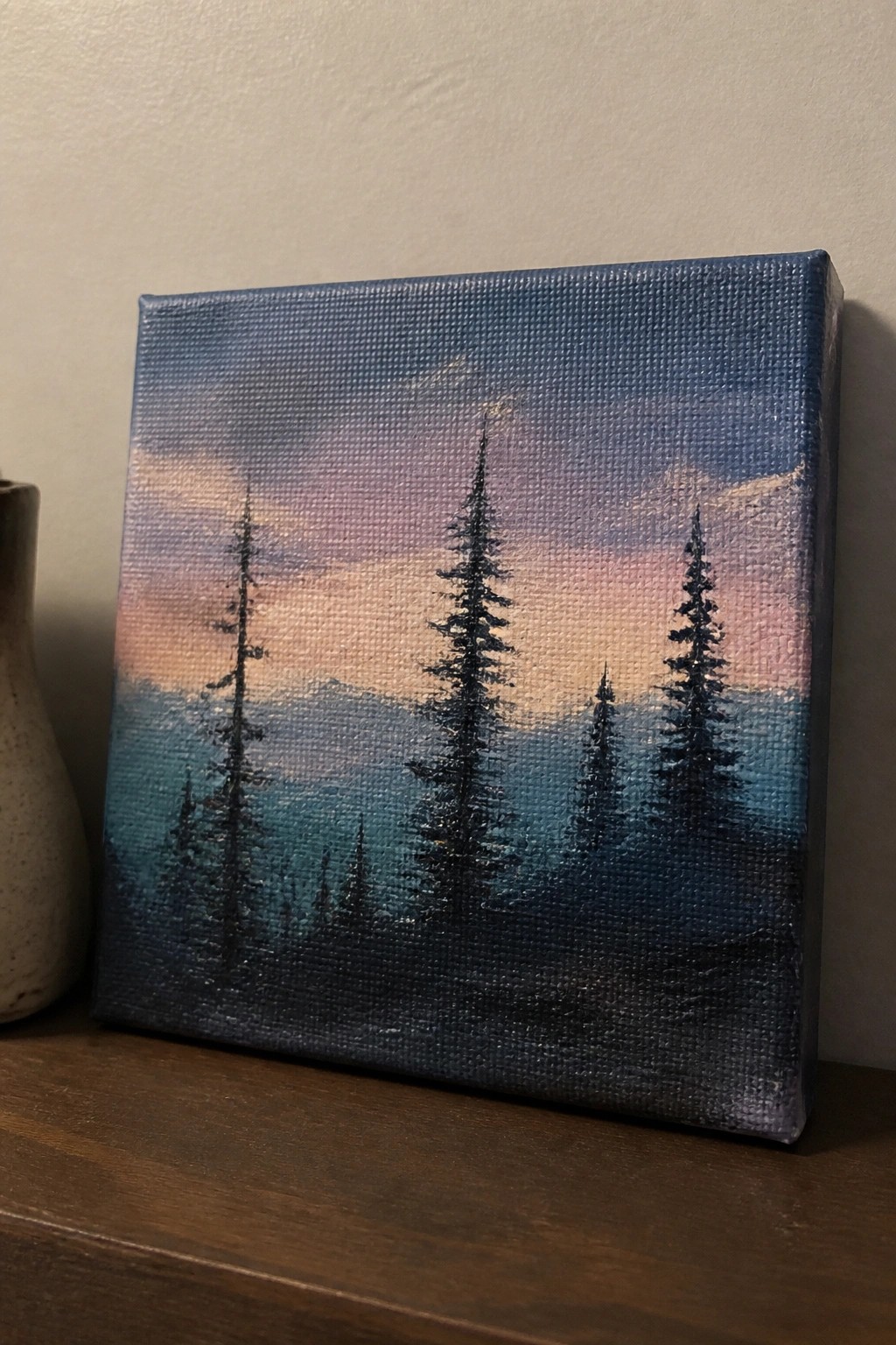 Small canvas painting of silhouetted pines against a pink, purple, and blue sky on wood.