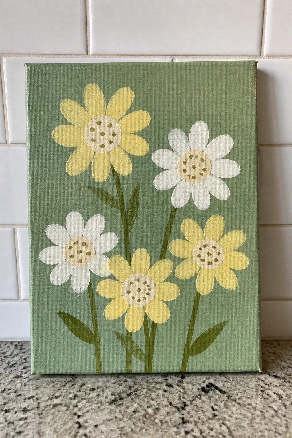 Green canvas painting of yellow and white daisies with green stems against white tiles