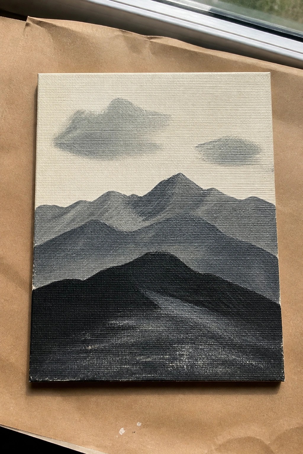 Layered grayscale mountain landscape painting on canvas atop brown paper