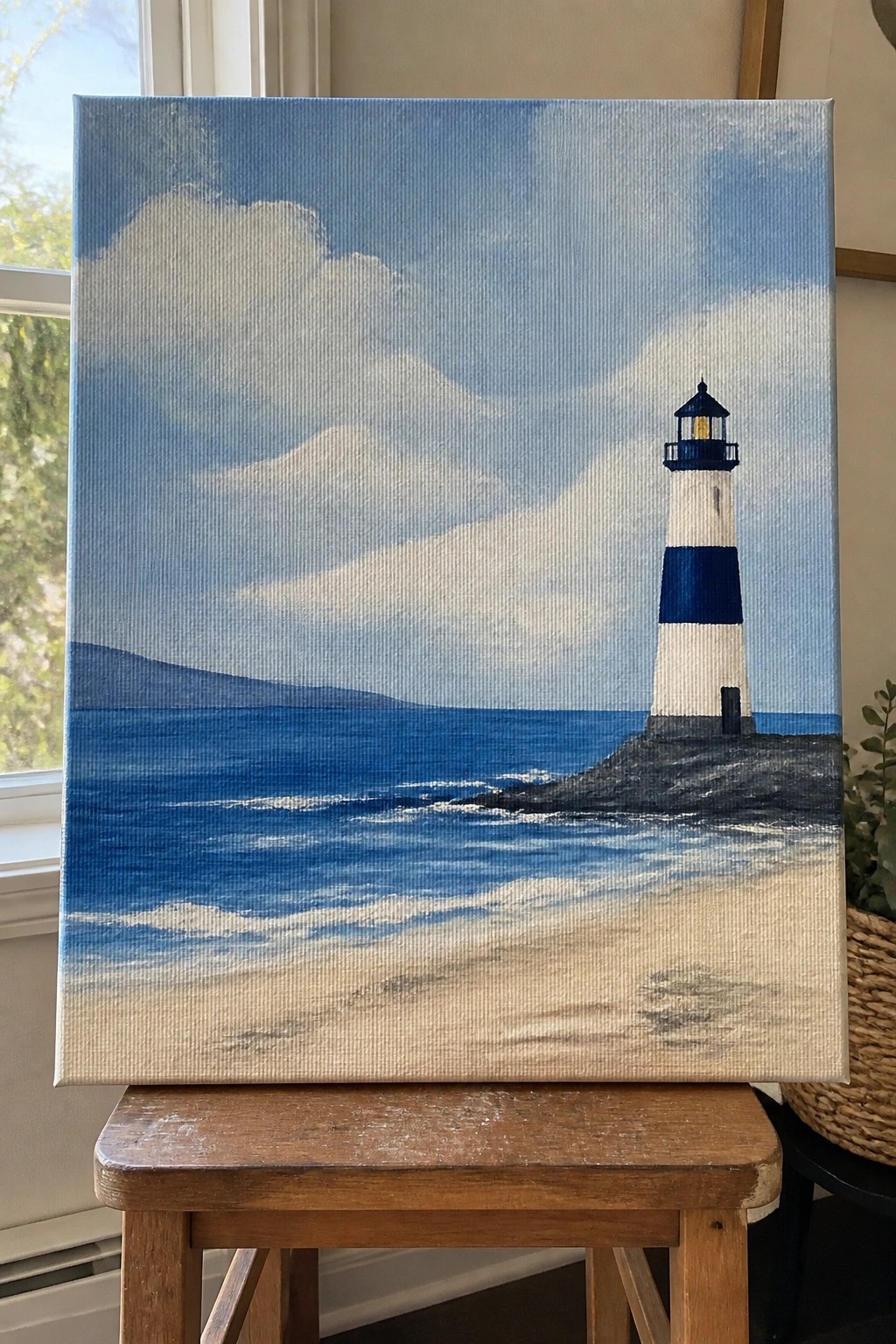 An acrylic painting of a white and blue striped lighthouse on a rocky shore next to a blue ocean with waves and a sandy beach in the foreground under a blue sky with clouds.