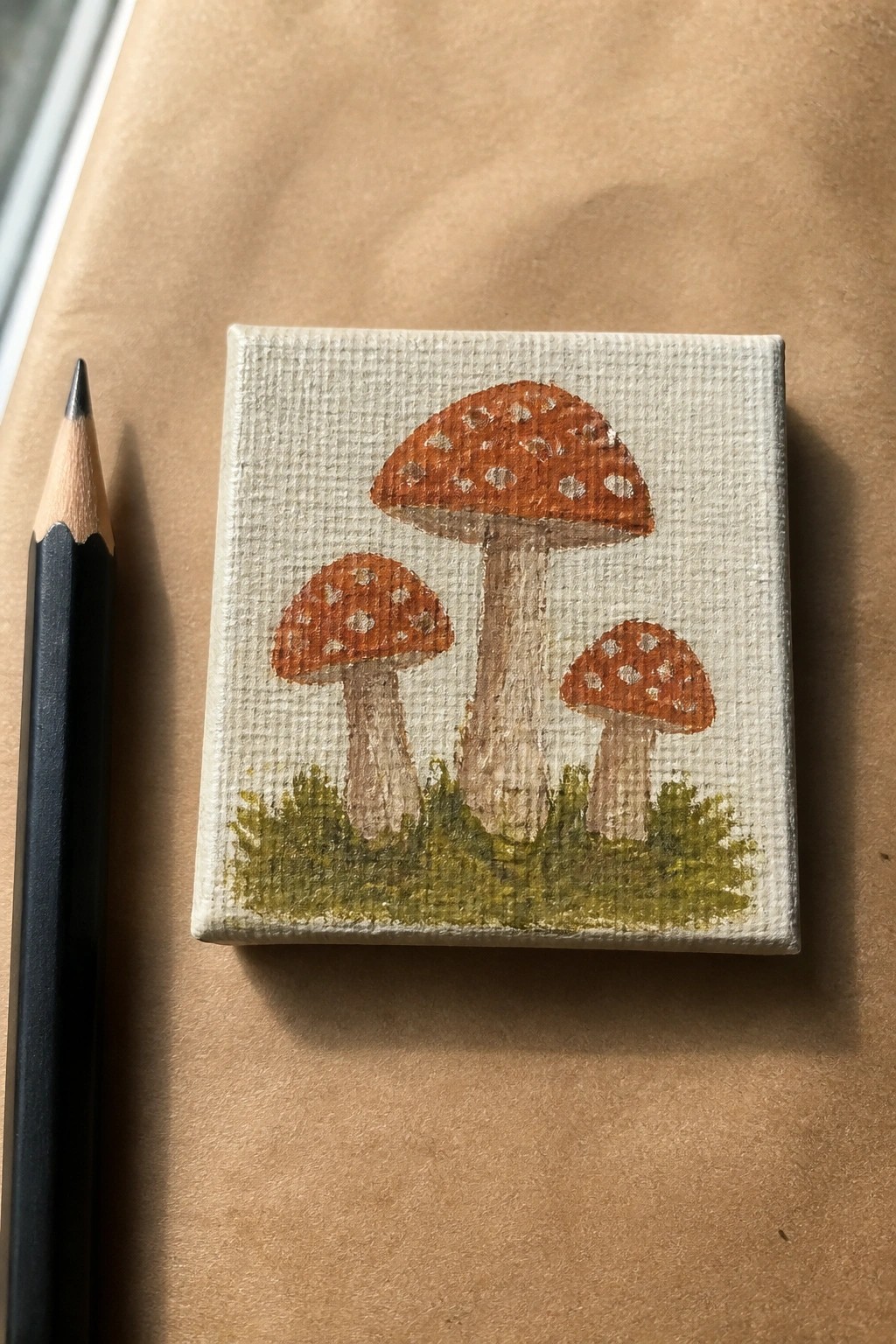 Mini canvas painting of three orange mushrooms with white spots beside a pencil