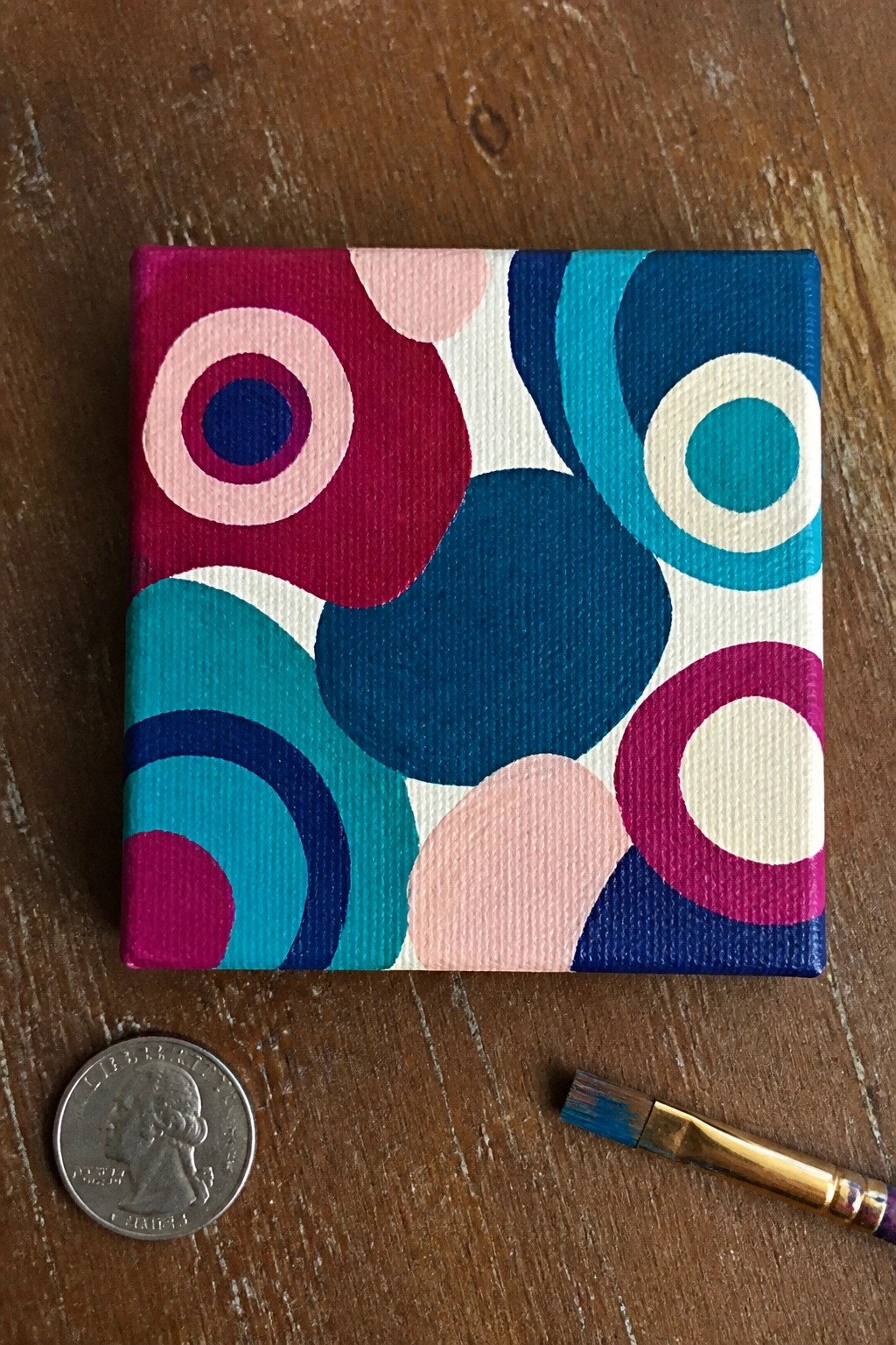Small canvas with colorful overlapping circles beside quarter and paintbrush on wood