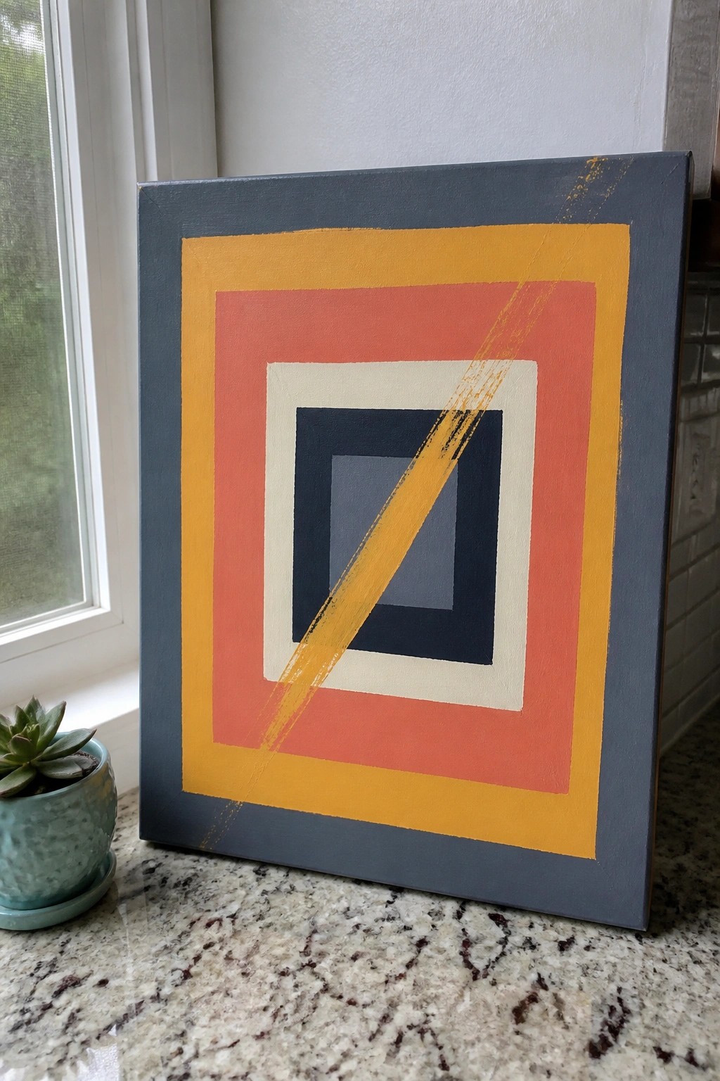 Colorful geometric painting with nested squares and yellow streak beside succulent on marble counter.