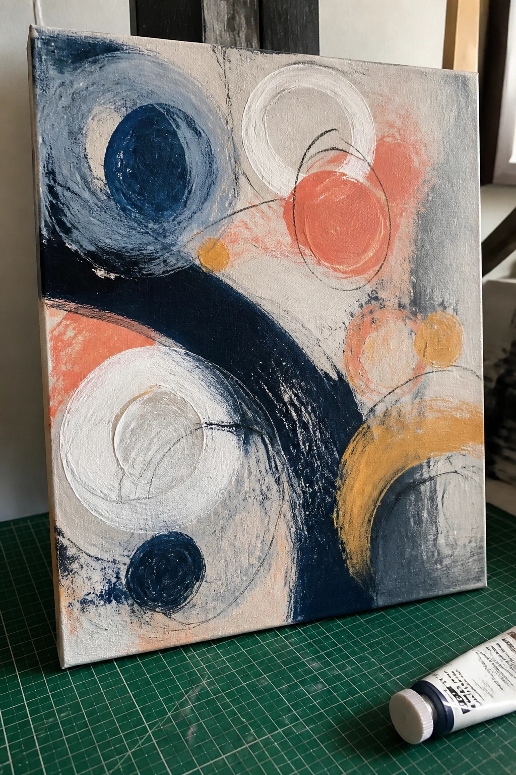 Abstract canvas painting with blue, coral, white, and yellow circles beside paint tube on green mat.