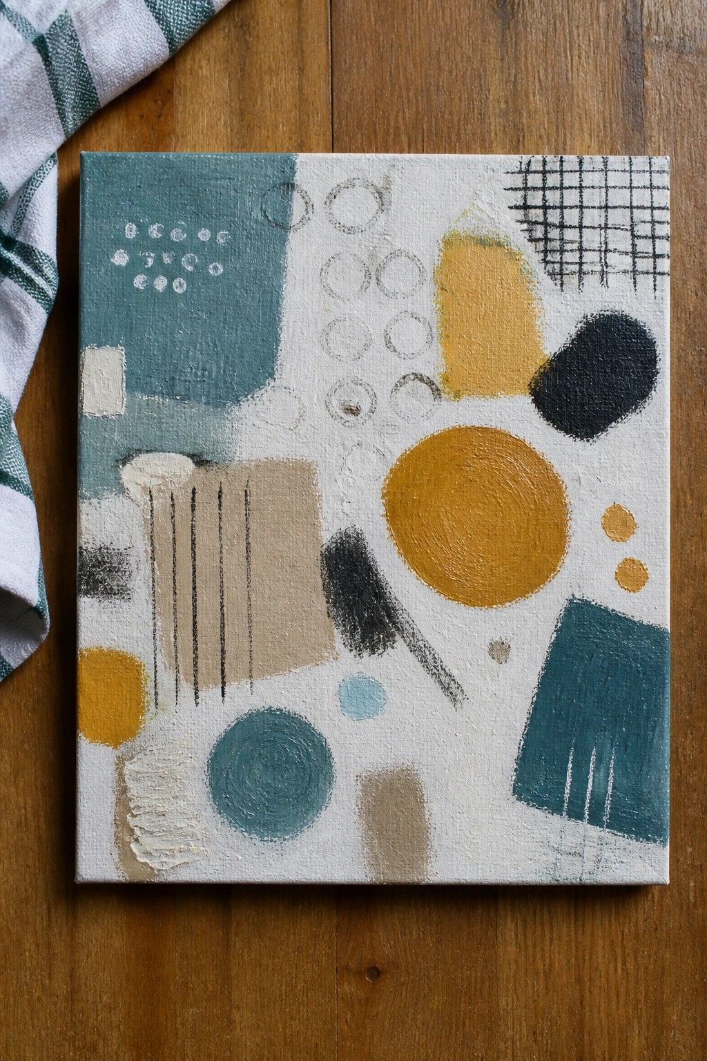 Abstract painting with teal, yellow, and black shapes on wooden surface beside checkered cloth