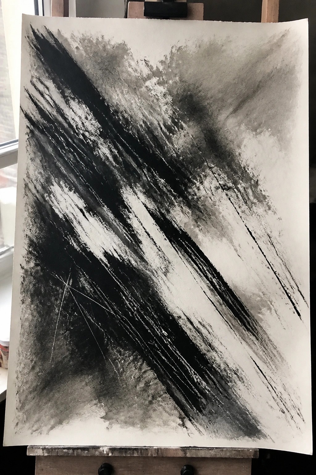 Bold black diagonal brushstrokes with gray washes fill a large white sheet of paper on a wooden easel.