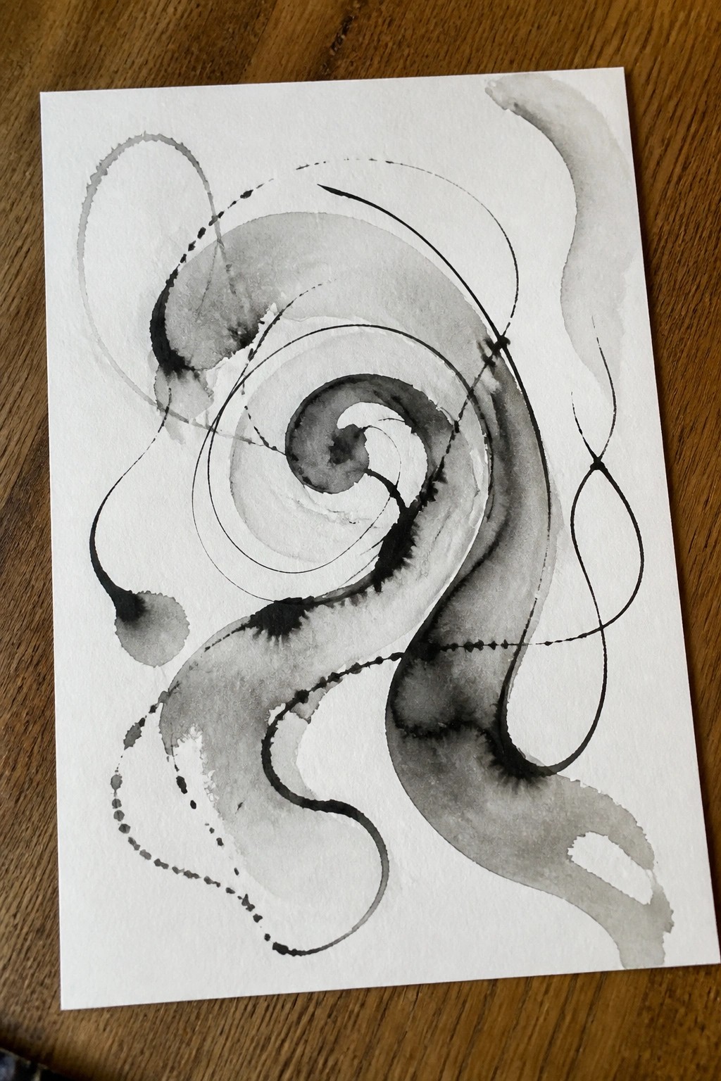 Abstract grayscale watercolor with swirling spirals and dotted lines on white paper atop wood.