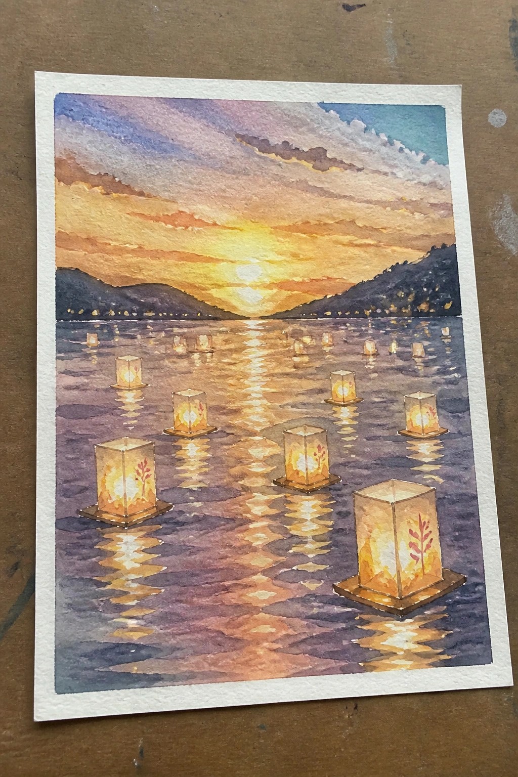 Watercolor of glowing lanterns floating on rippling water beneath orange sunset sky and mountains.